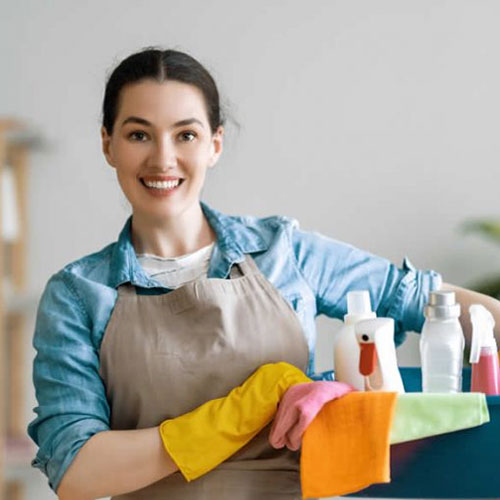 A smiling woman in an apron and rubber gloves holding a caddy filled with cleaning supplies, ready for house cleaning.