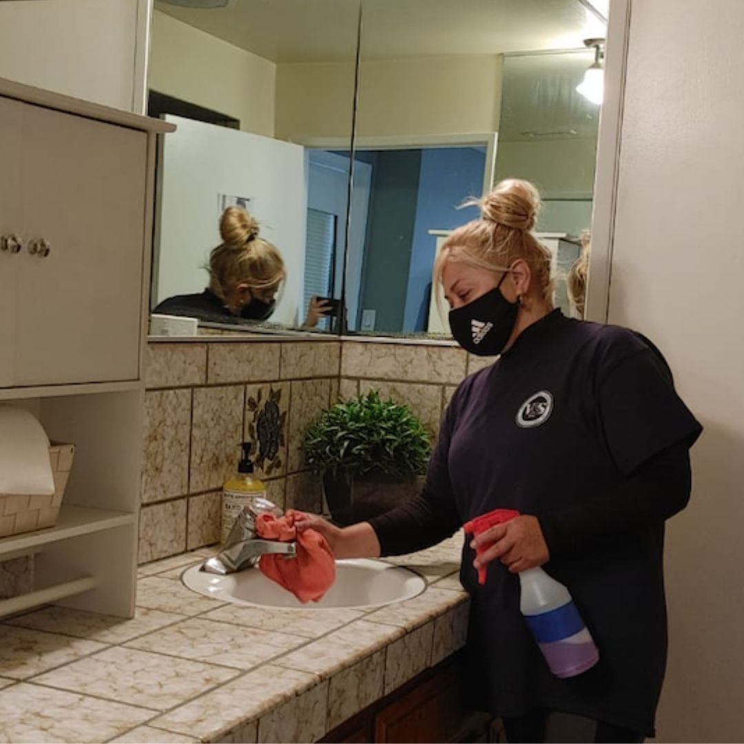 A woman wearing a face mask and cleaning a bathroom sink with a cloth and spray bottle, ensuring hygiene and cleanliness.