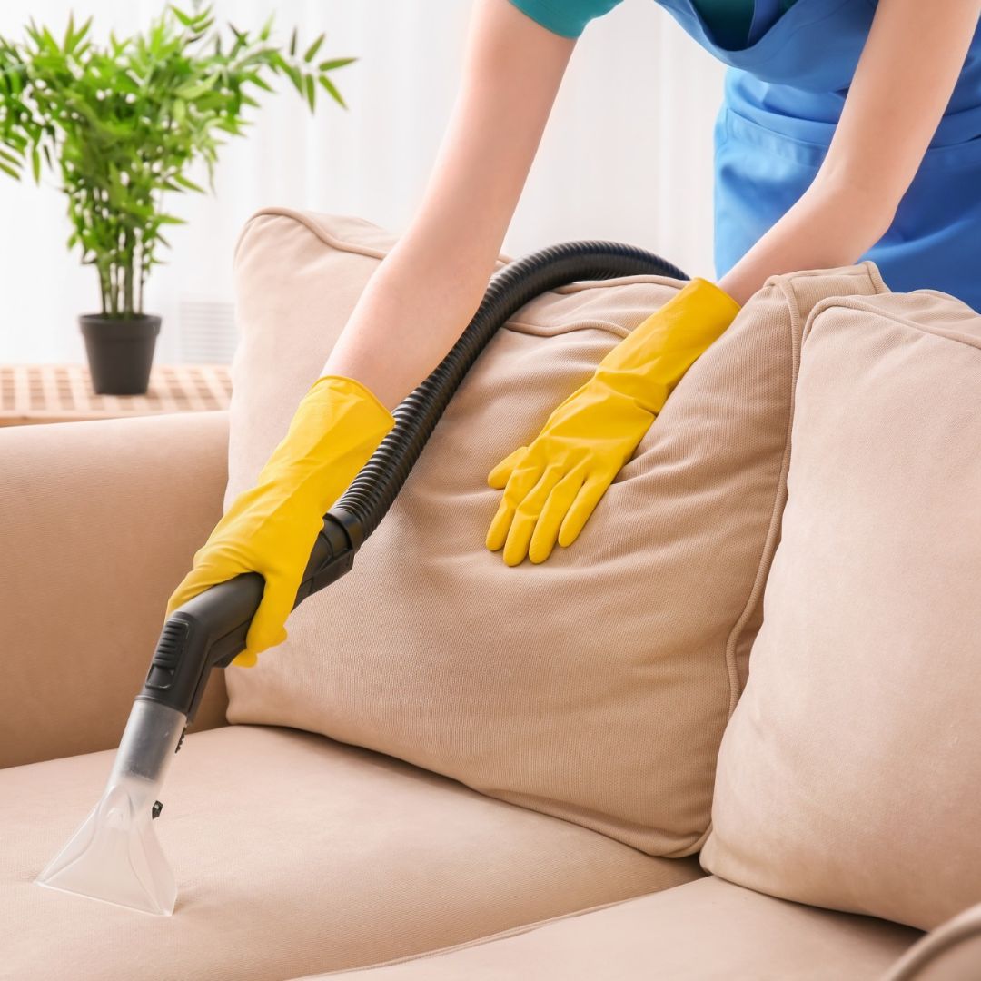 A close-up of a person using an upholstery cleaning machine on a beige sofa, wearing yellow rubber gloves to protect their hands.